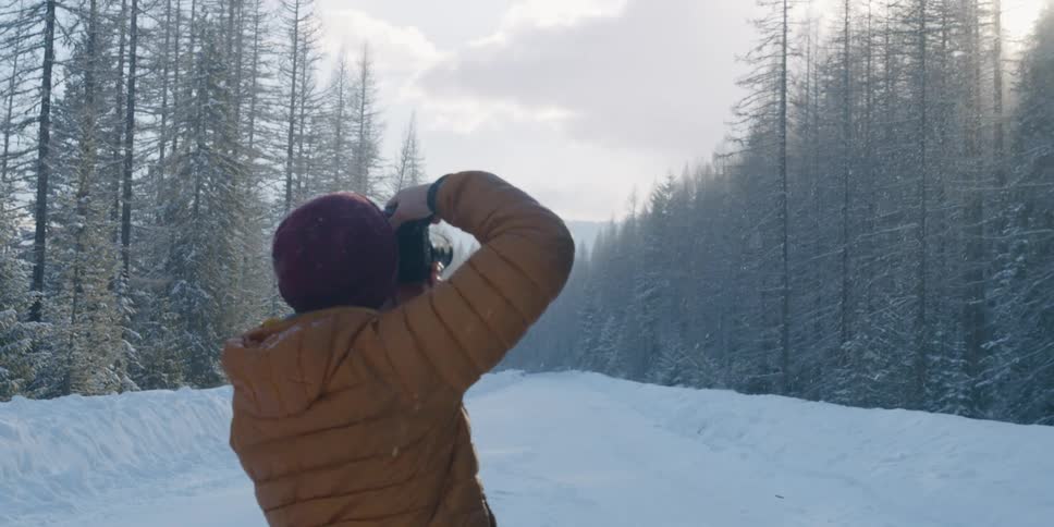 A person with a hat and coat taking a picture with a camera with trees the sky and clouds in the background