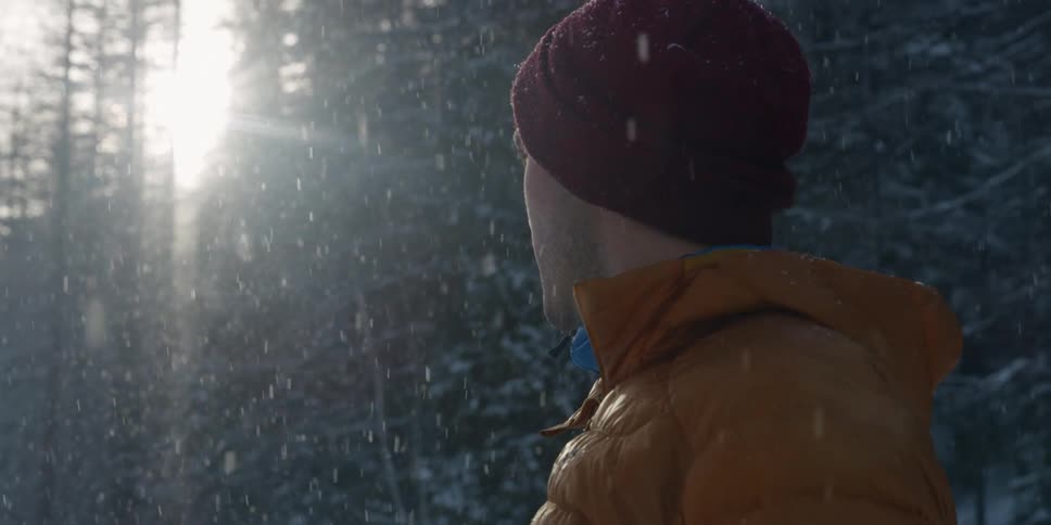 A man wears an orange parka in the forest and stands still as snow falls around him