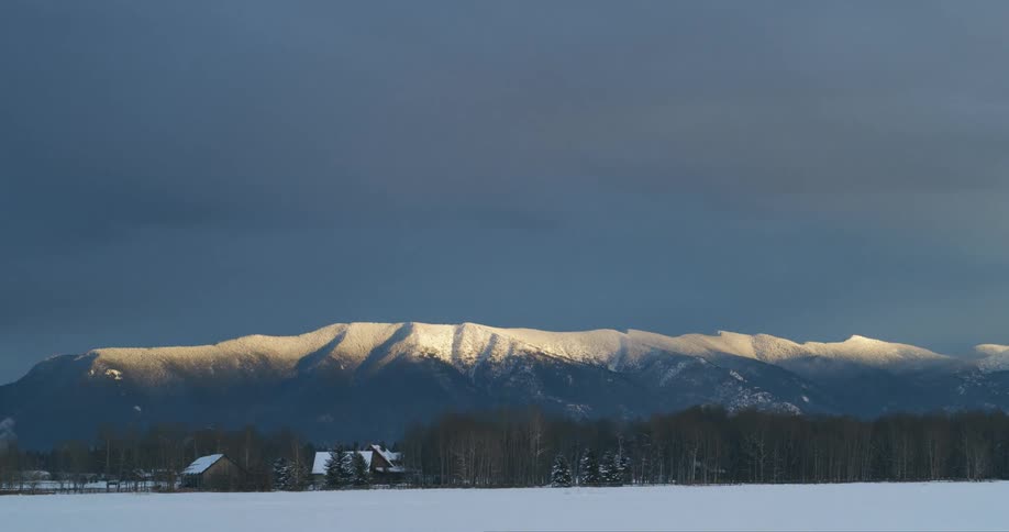A snowy field with cabins and trees with a mountain range the sky and clouds in the background