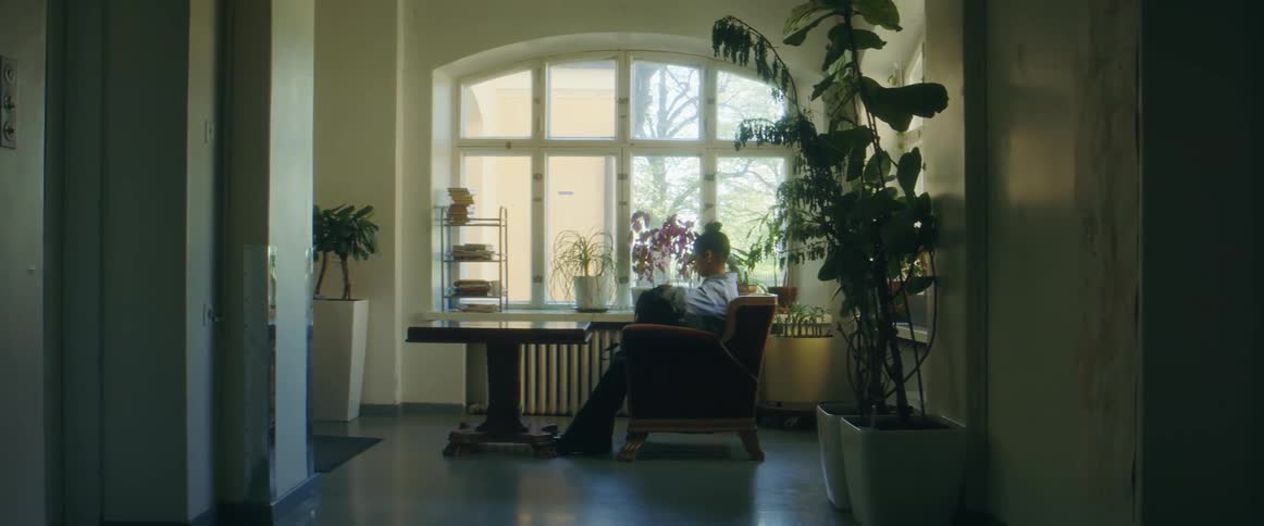 A woman walks down a hallway and sits in a comfortable chair next to a large window