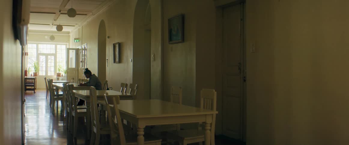 A female sits at a table in a hall with other tables and writes