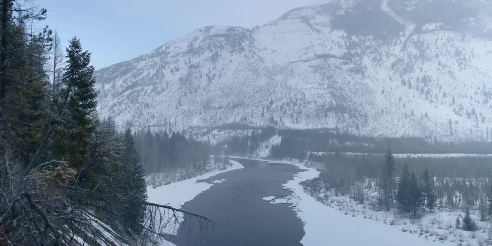 Snow falls over a rural highway that curves past mountains