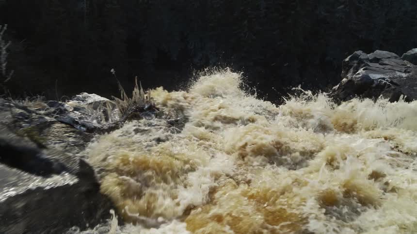 Foaming water churns turning brown with dirt as it passes ice crusted vegetation before flowing down waterfall
