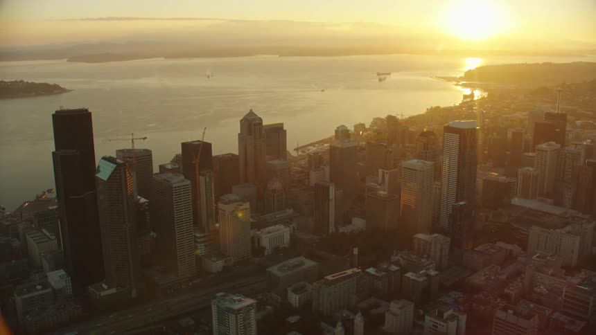 Sun shines from red sky reflecting off water near shipping vessels as traffic flows along freeway through city center