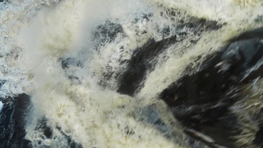 Water churns producing brown tinted foam then flows down black rock past white ice