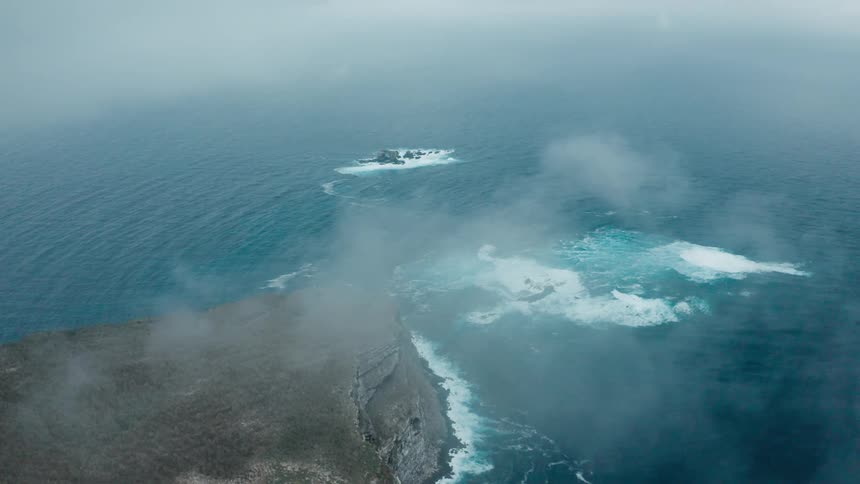 Waves break and splash against rocks and cliff as clouds and fog move over