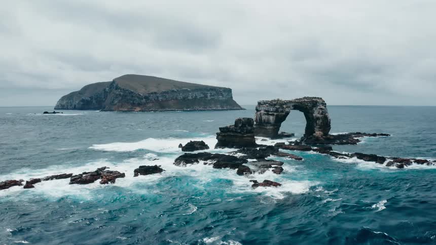 Waves splashing against a natural arch and rocks in the ocean with an island in the distance