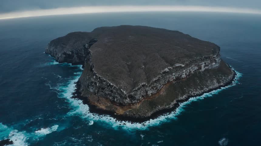 A rocky island with high cliffs and a very small beach area sits in the middle of a large body of water with waves crashing against it