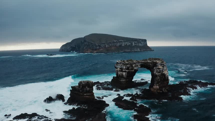 Waves crashes against island and worn away islet arch in middle of blue ocean under overcast sky as birds fly overhead