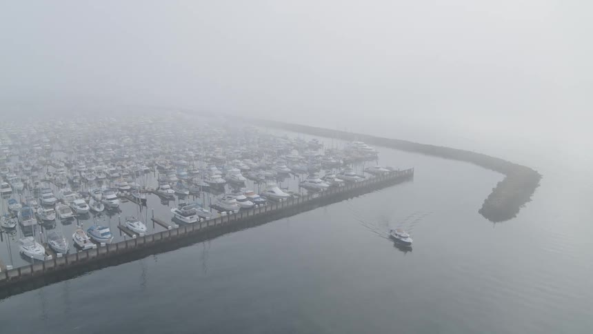 A motor boat departs from a marina around a breakwater as dense fog moves in