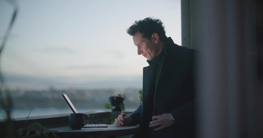 Man with a black coat sitting at table drinks a cup of coffee while making a video call on tablet at dusk