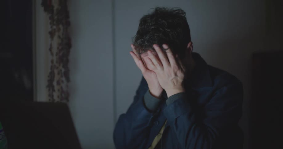 A man sits at a desk with a laptop and rubs his face with exhaustion