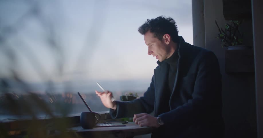 Man sits at a table next to a window and talks to someone on his laptop