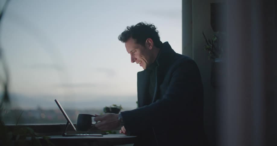 Man with black coat sitting at table and drinks a cup of coffee while making video call on tablet