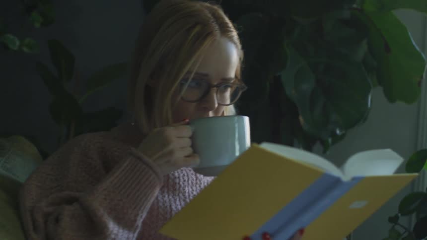 Young woman drinks a hot beverage while reading a book in the couch at home