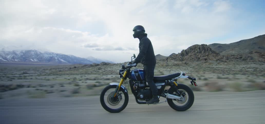 A person with a helmet riding a motorcycle with a desert a mountain range the sky and clouds in the background