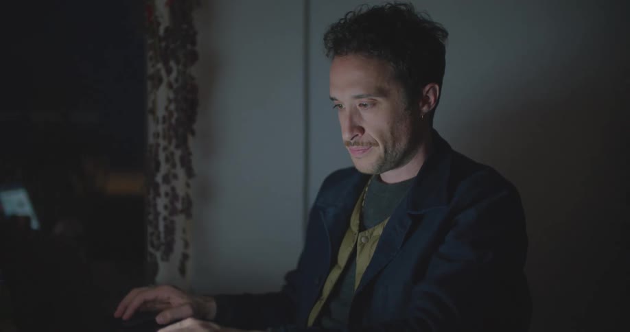 A male is sitting in a dark room typing on a laptop