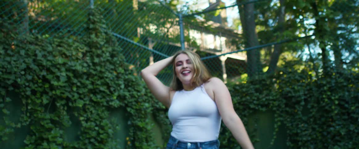 Blonde woman dances next to climbing vine covered walled topped by chain link fence near large homes