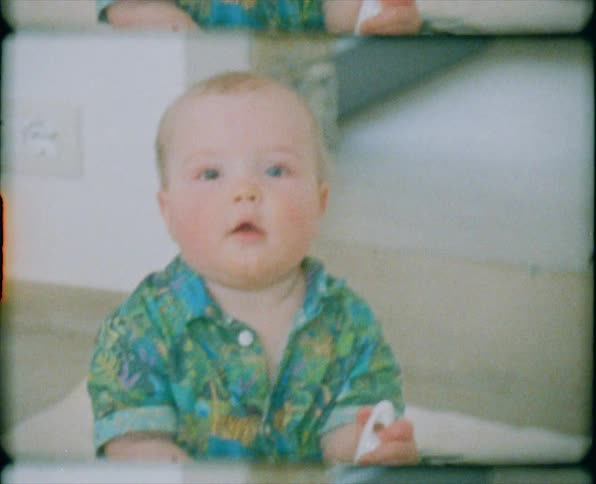 A baby boy in a shirt and diaper sits on a blanket in a carpeted room