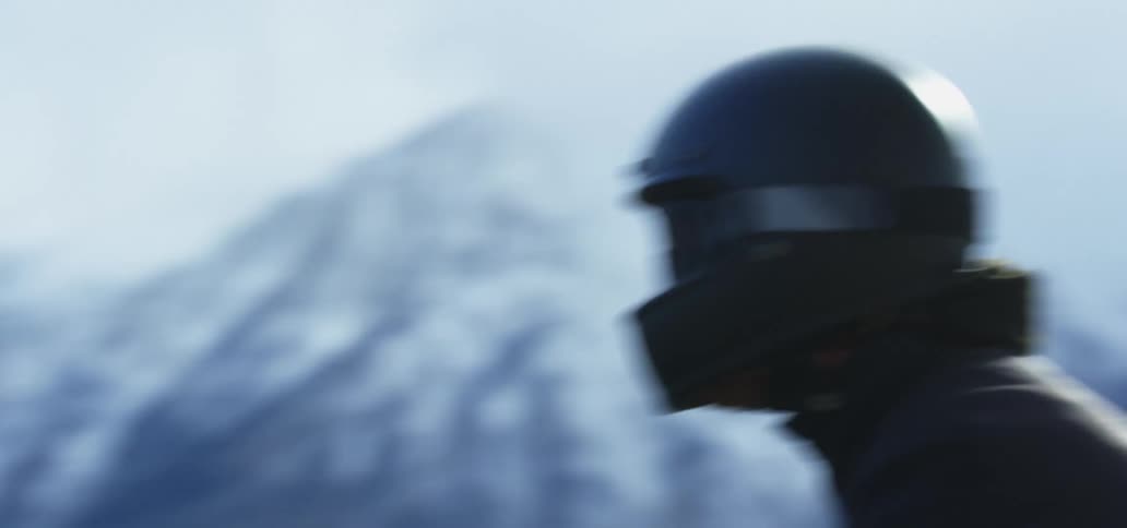 Person wearing a crash helmet riding a motorcycle on a rural road near the snow covered mountains
