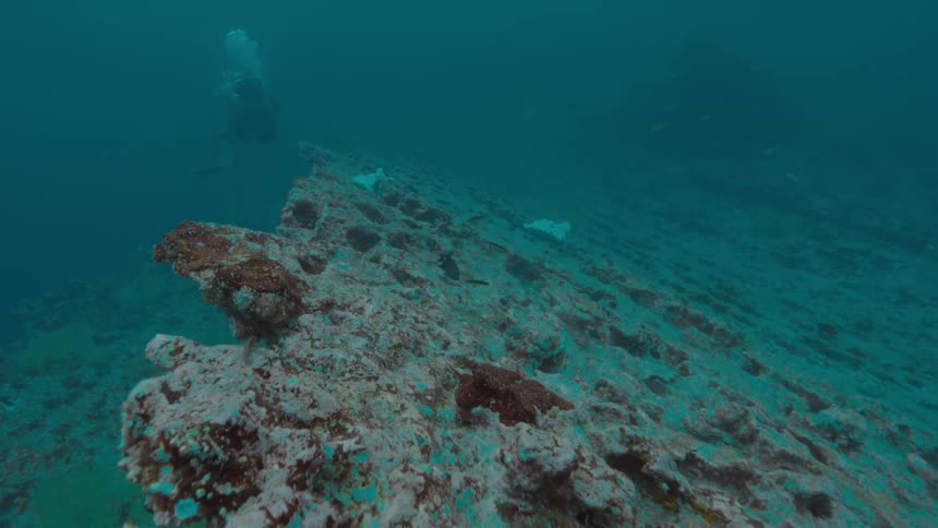 Scuba diver releases bubbles while swimming near sloping reef overhang in murky water