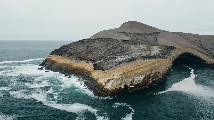 Waves crash against lands with sheer cliff and sea cave under overcast sky on misty day