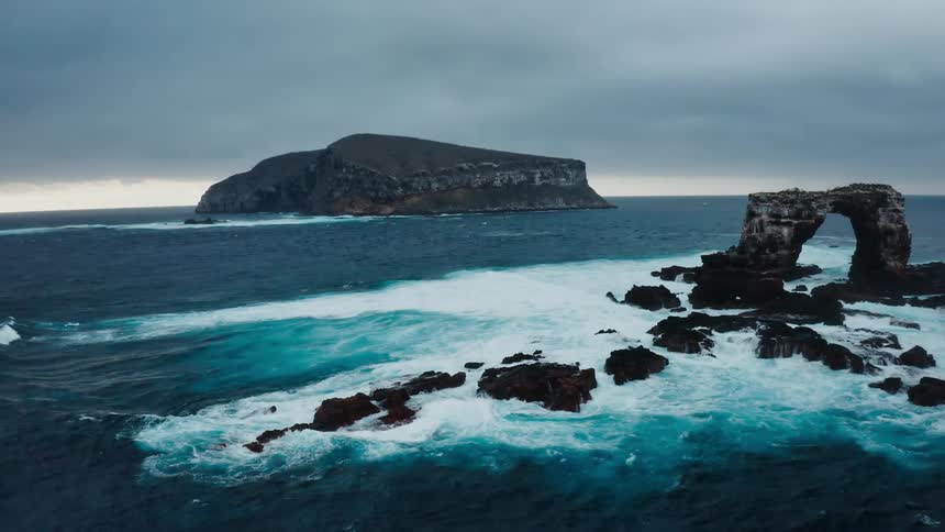 Waves splashing against a natural arch and rocks in the ocean with an island in the distance