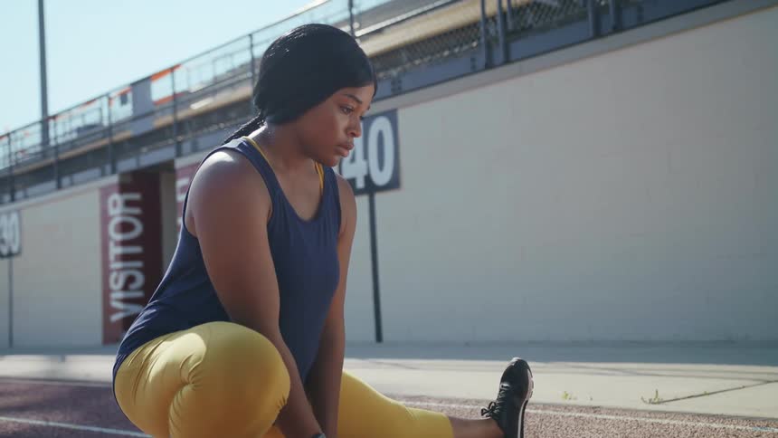 Black woman stretches her leg while crouched on an outdoor track