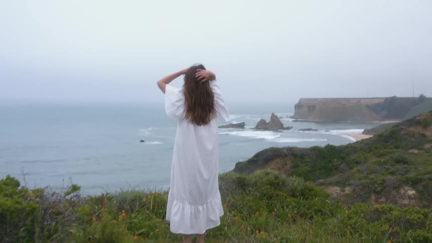 Woman in a long dress stands on a hill and looks out at the ocean