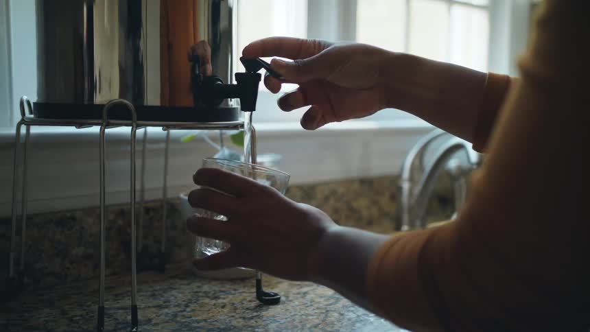 A woman gets a glass for the kitchen cabinet and fills it with water from a container on the counter