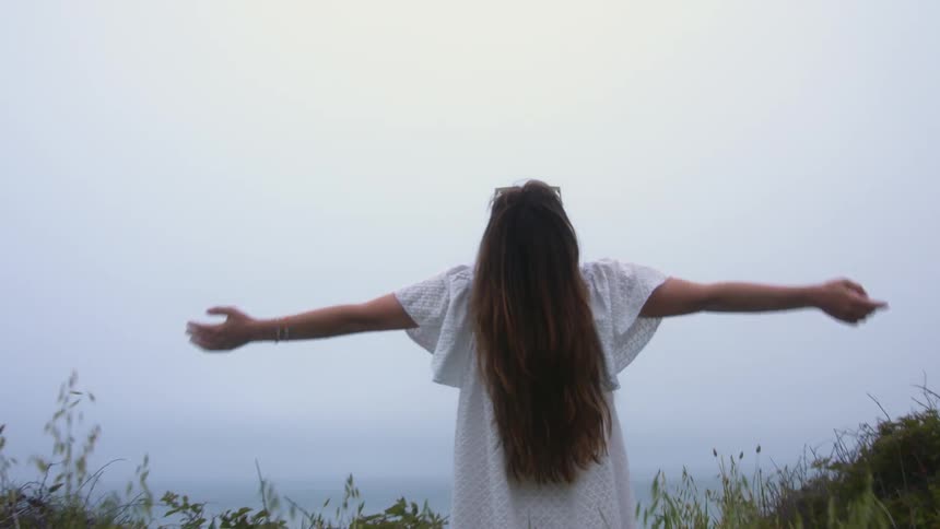 A woman standing and stretching on a grassy field near the coast with the ocean sky and clouds in the background