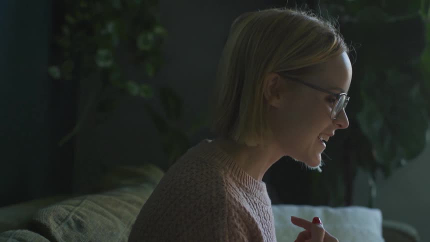 Blonde woman with glasses and a pink knitted sweater makes a video call on smartphone in the living room at home