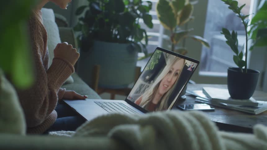 Blonde woman with pink knitted sweater sitting on couch makes video call on macbook pro laptop in the living room at home