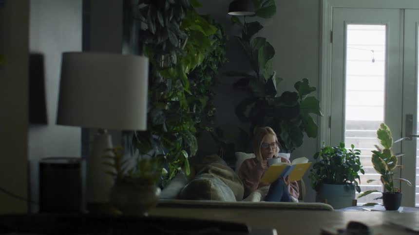 Woman wearing sweater reclines on couch sipping from mug as she reads surrounded by houseplants