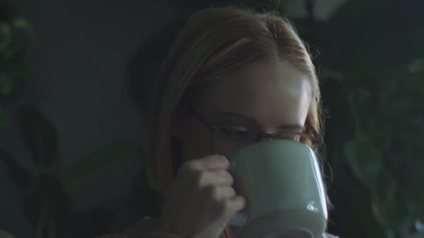 Young blonde woman wearing glasses blows a hot beverage while sitting at home