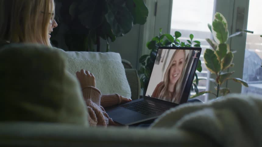 Woman wearing sweater sitting on couch converses via video call on laptop with a another woman