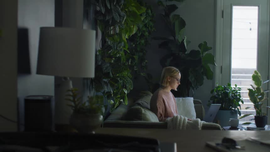 A woman holding her laptop sits down on a couch then settles in