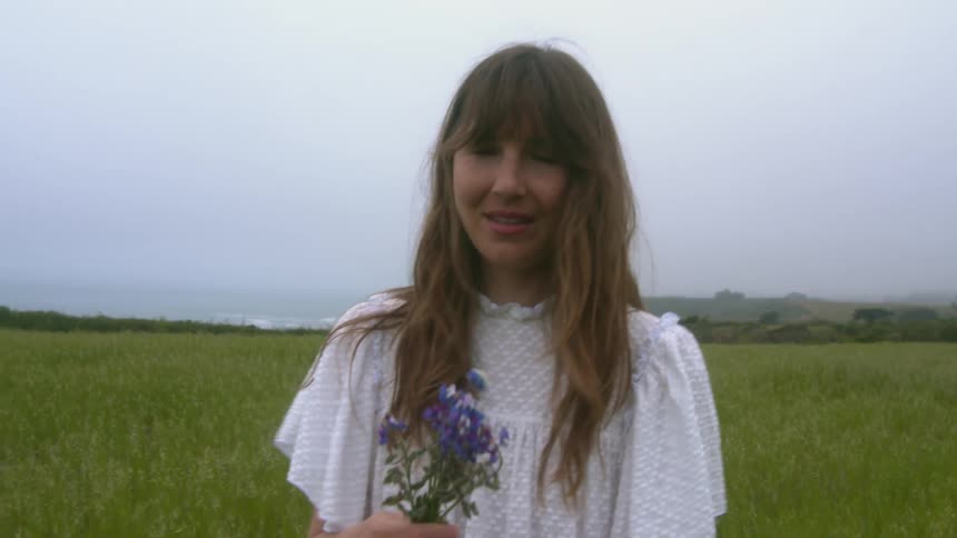 A woman holding flowers and walking on a grassy field with the sky and clouds in the background