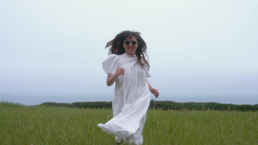 Woman with sunglasses and white dress runs in grassy field on a cloudy day