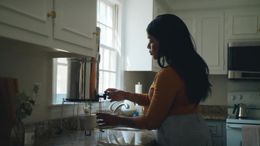 A woman gets a class out of the cabinet and fills it with water from a container on the kitchen counter