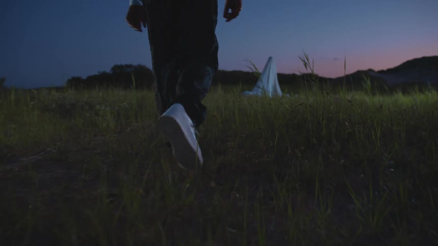 A person walking on grass towards another person covered in a sheet with the sky and clouds in the background