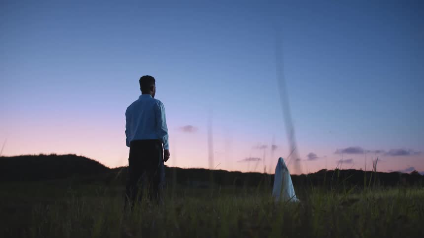 Man stands in field facing shrouded figurebacked by red tinted horizon and forest