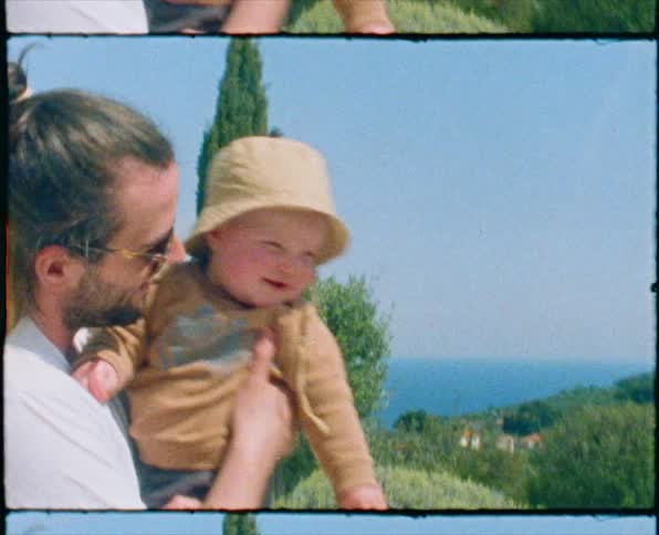 A man with sunglasses holding a baby with trees the ocean sky and clouds in the background