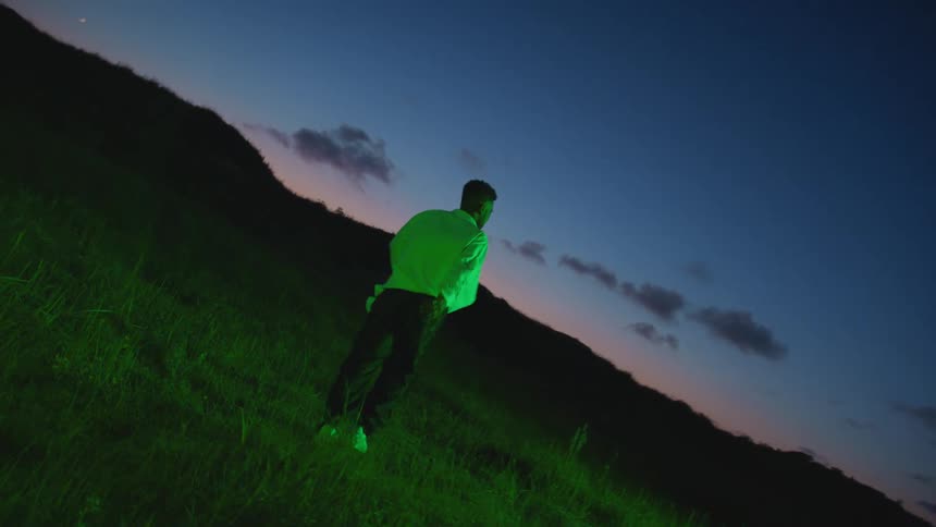 A man walking on a grassy field with hills the sky and clouds in the background