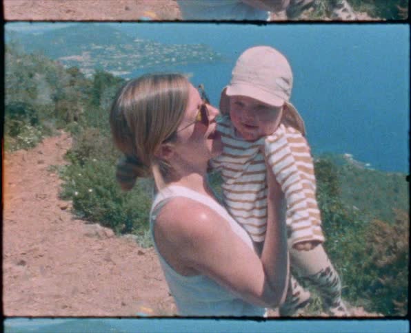 Mother carries and kisses toddler standing on dirt trail next to water with town in distance