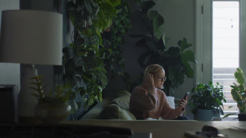Woman sits on couch in room decorated with many houseplants working on mobile phone