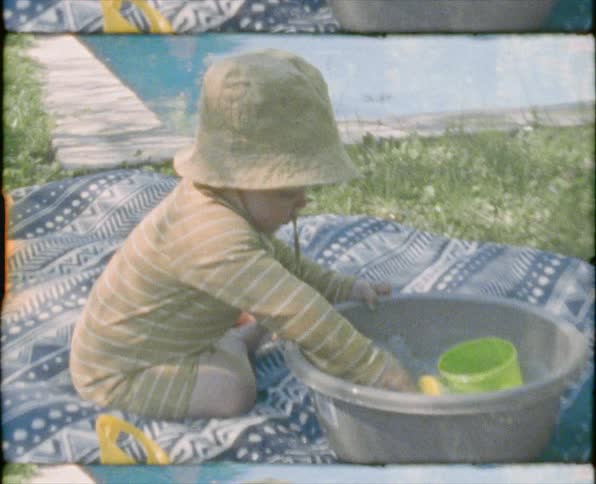 A baby boy playing in a bowl of water with a pool and a woman sunbathing in the background