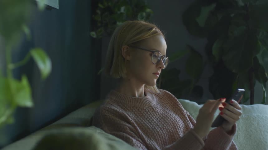 A woman sits on a couch scrolling on her cell phone