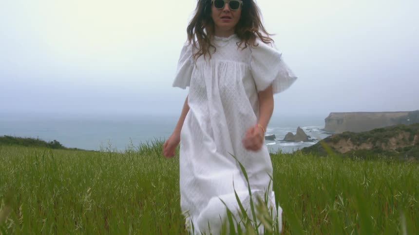 A woman with sunglasses jogging on a grassy field near the coast with the ocean sky and clouds in the background
