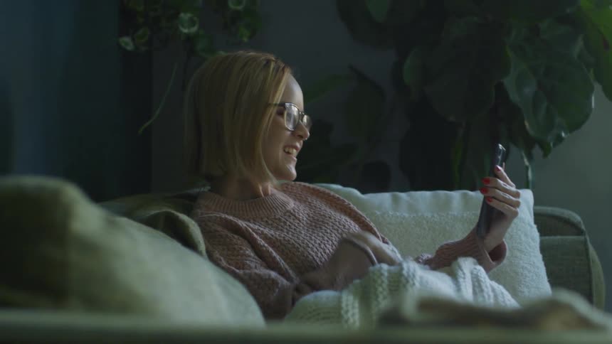Blonde woman with glasses and a pink sweater holds smartphone and makes a video call in the living room at home